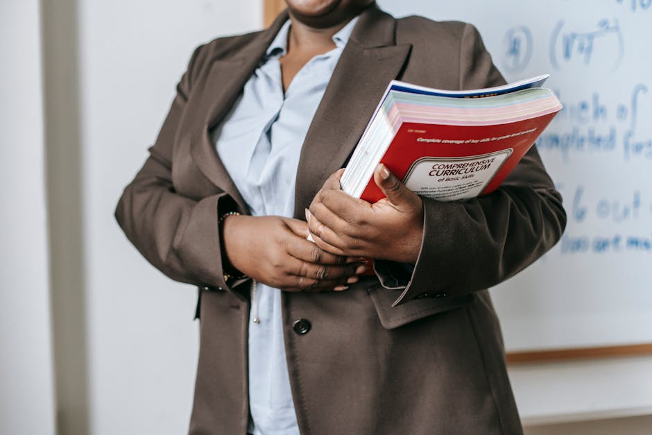 Crop faceless African American female teacher in formal clothes standing near whiteboard with textbooks in hand during lesson in classroom